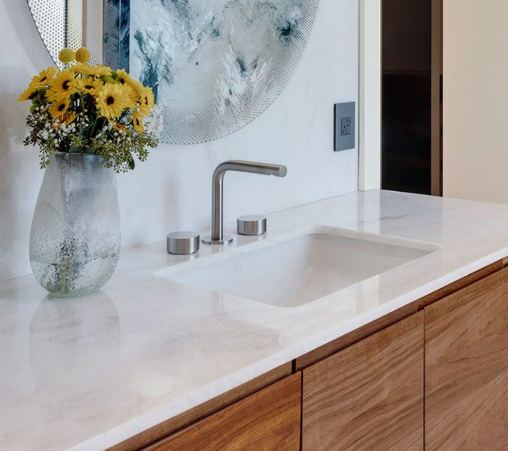 Modern bathroom sink with a chrome faucet on a marble countertop, wooden cabinets below, and a vase of yellow and white flowers next to a round, textured mirror.