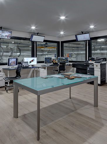 Modern office with multiple desks, computers, and chairs. Large windows reveal an industrial setting outside. Central glass table on a wooden floor.