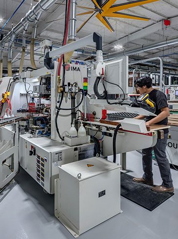 A person operating a large industrial machine in a factory setting, surrounded by panels and machinery, with overhead pipes and lighting visible.