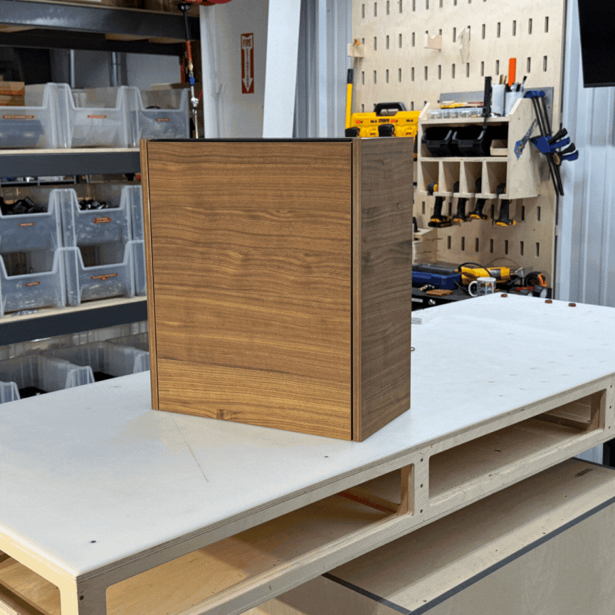 A wooden cabinet box sits on a workbench in a workshop, with tools and storage bins visible in the background.