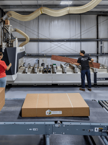 Two workers operate a large industrial machine in a factory, with cardboard boxes on a conveyor belt in the foreground.