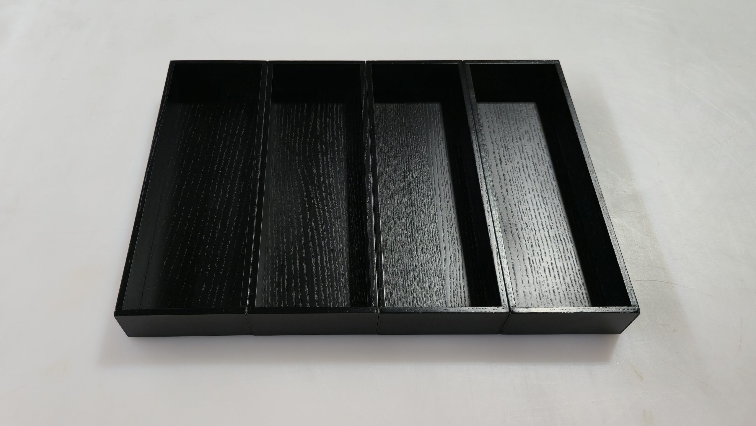 Four empty, rectangular black wooden trays are arranged side by side on a white surface.