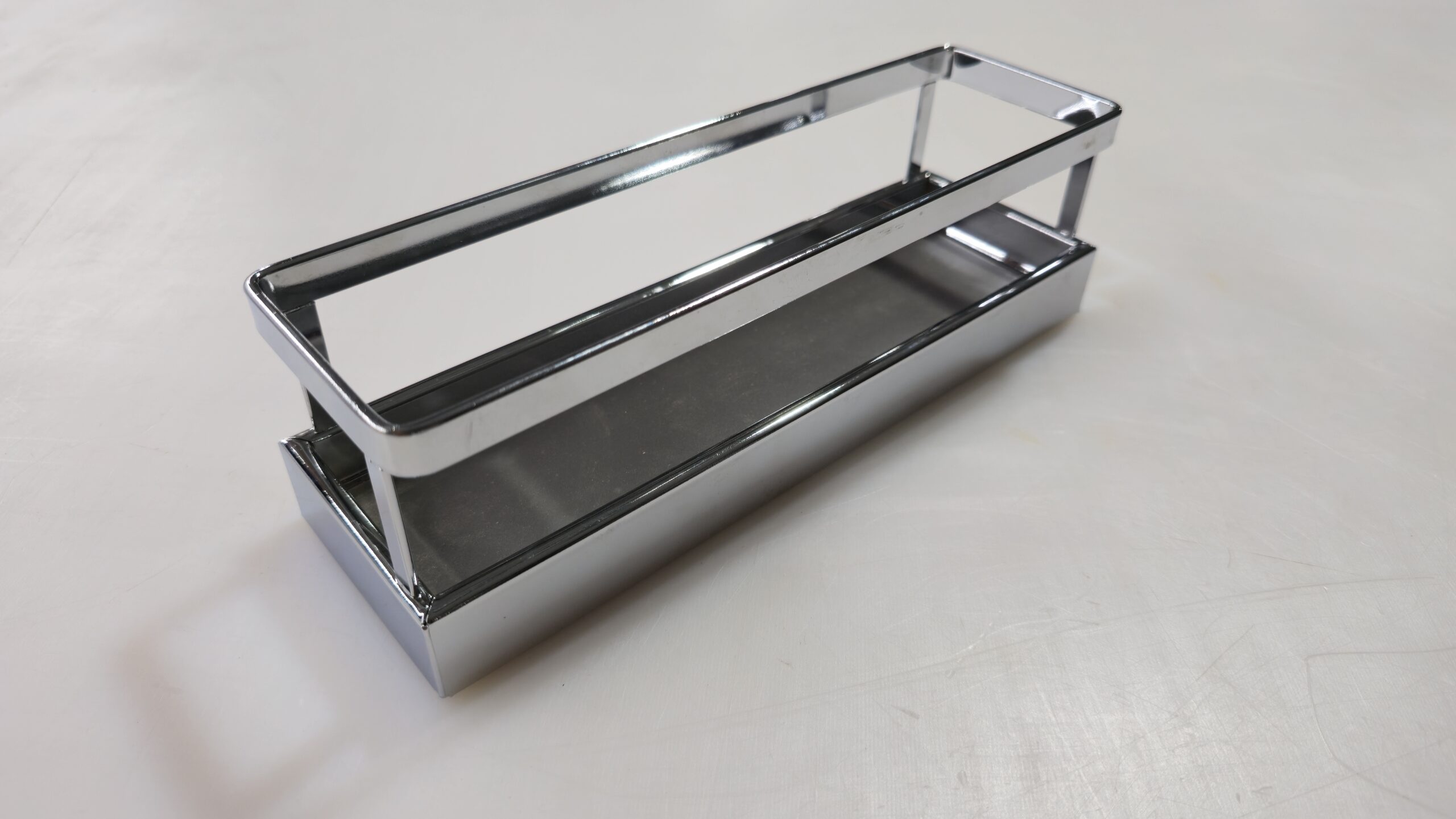 A rectangular, chrome-finished metal shelf with open sides and a flat base, placed on a light-colored surface.