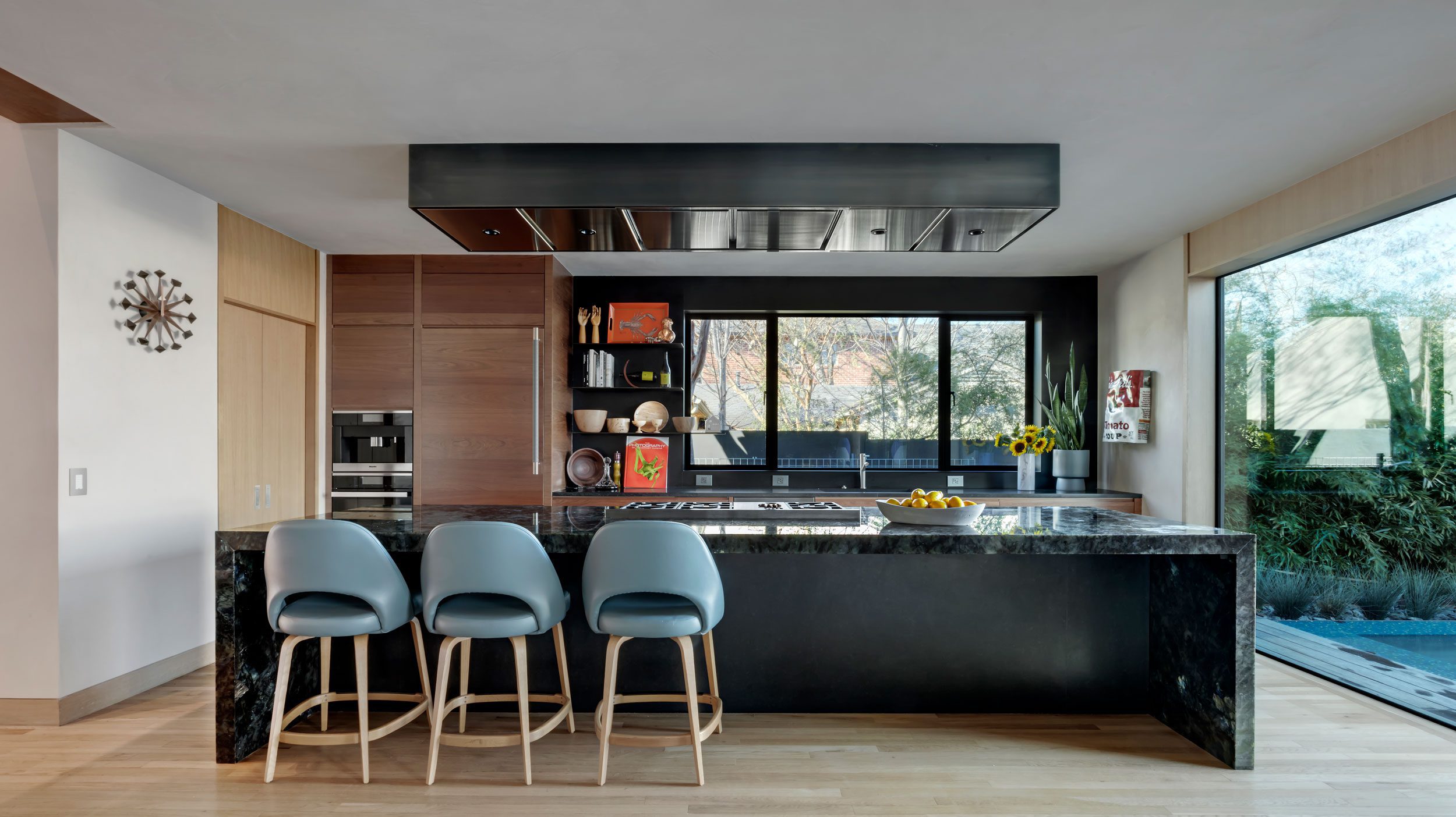 A modern kitchen with a black marble island, three blue barstools, built-in wooden cabinets, and large windows overlooking greenery.