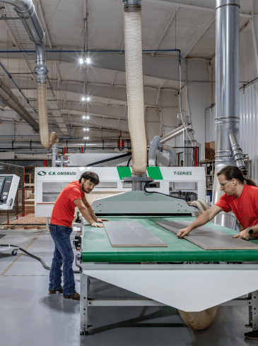 Three people work in a factory, operating machinery and handling large panels on an assembly line in a brightly lit industrial setting.