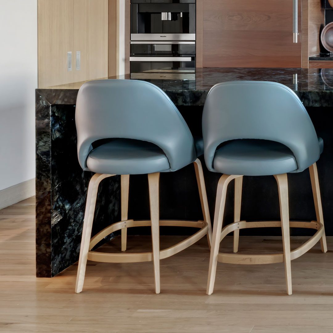 Two modern blue barstools with wooden legs sit at a dark marble kitchen counter with built-in cabinets and appliances in the background.