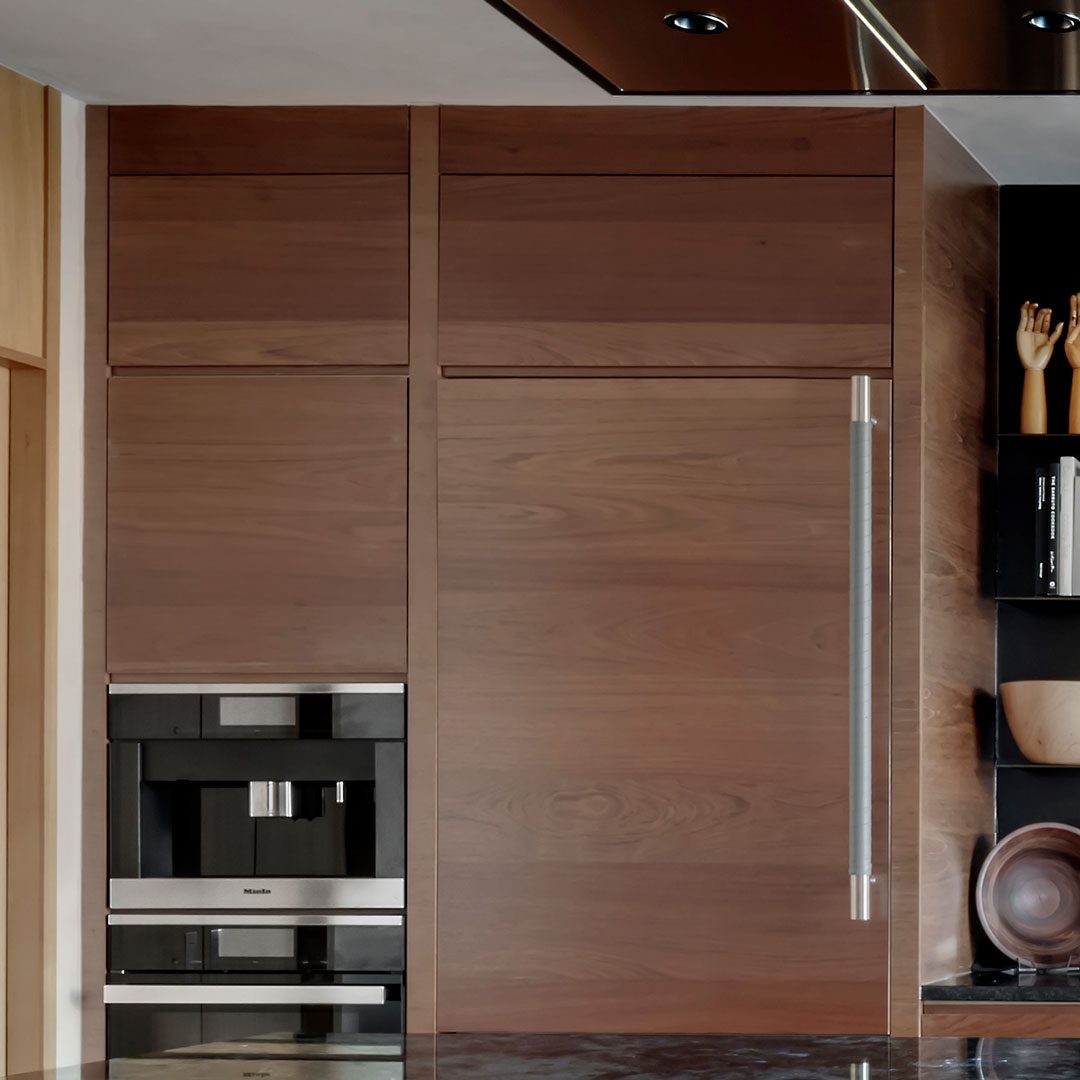 Built-in stainless steel coffee machine and refrigerator with wood paneling, adjacent to shelves holding decorative items and bowls.