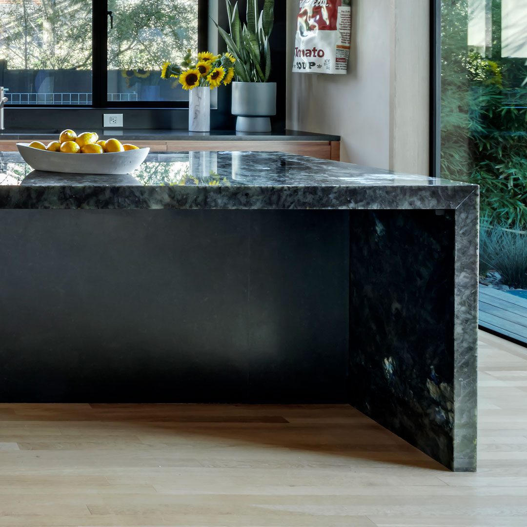 A modern kitchen features a dark marble island with a bowl of lemons, sunflowers in a vase, and large windows overlooking greenery.