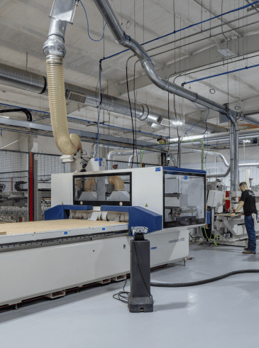 Two people operate large woodworking machinery in a clean, industrial facility with metal ducts and safety barriers.