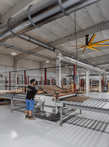 A worker operates a machine to process large sheets of material in a spacious, modern factory with exposed ductwork and a yellow ceiling fan.