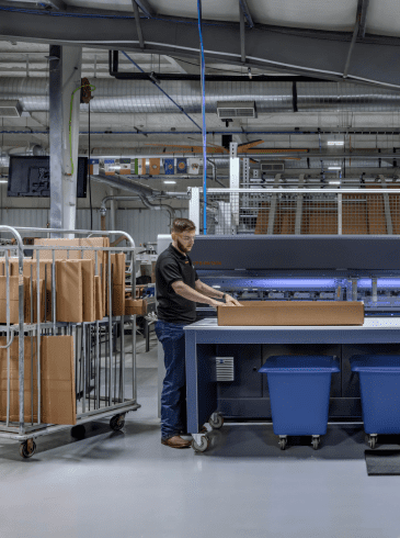 Two people operate a large packaging machine in a factory, assembling cardboard boxes with carts, bins, and computer monitors nearby.